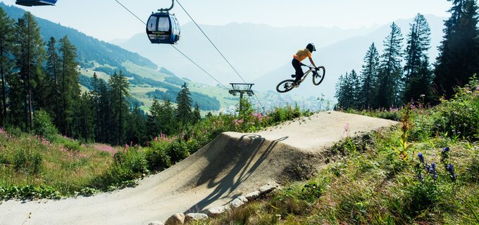 Ein Mountainbiker springt über einen Hügel, während eine Seilbahn im Hintergrund schwebt.