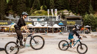 Zwei Mountainbiker fahren in einem Bikepark mit Bergblick.