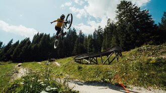 Ein Mountainbiker springt über einen Kicker in einer Berglandschaft.