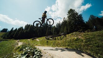 A mountain biker leaps into the air over a ramp, surrounded by trees and blue sky.
