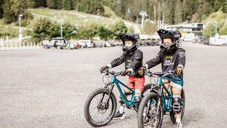 Two kids in protective gear riding mountain bikes in a parking lot.