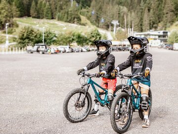 Zwei Kinder in Schutzausrüstung fahren Mountainbikes auf einem Parkplatz.