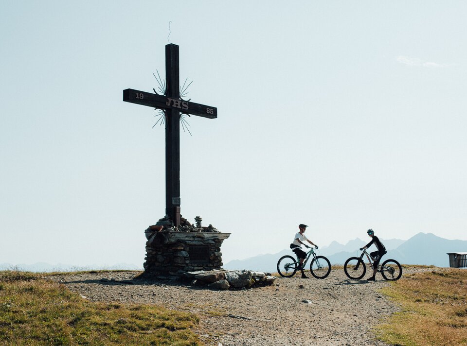 Two mountain bikers at a summit cross in the mountains.