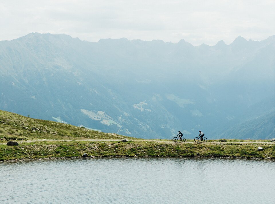 Two cyclists riding along a lake in the mountains.