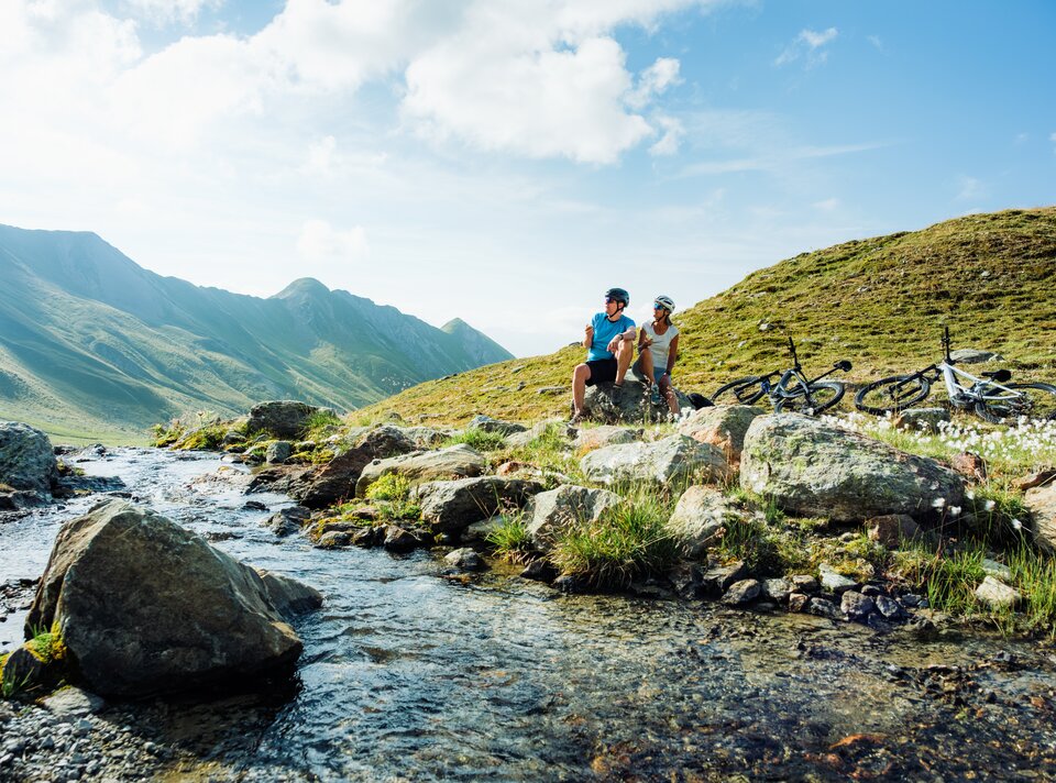 Two bikers relaxing by a clear mountain stream.