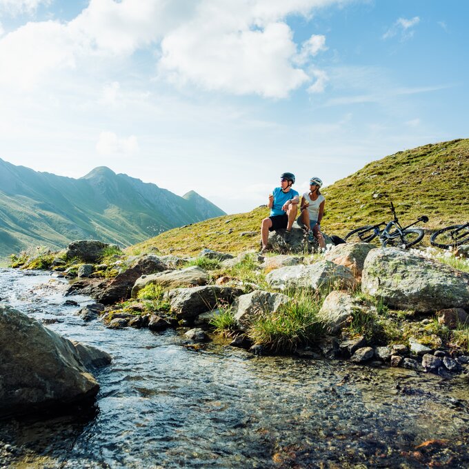 Two bikers relaxing by a clear mountain stream.