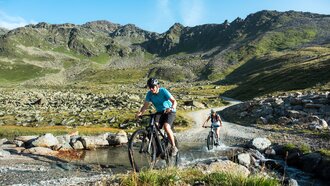 Two mountain bikers ride through a stream in the mountains.